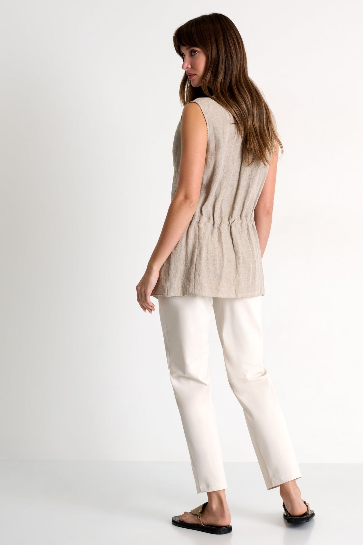Woman wearing a beige sleeveless top and white pants on a white background